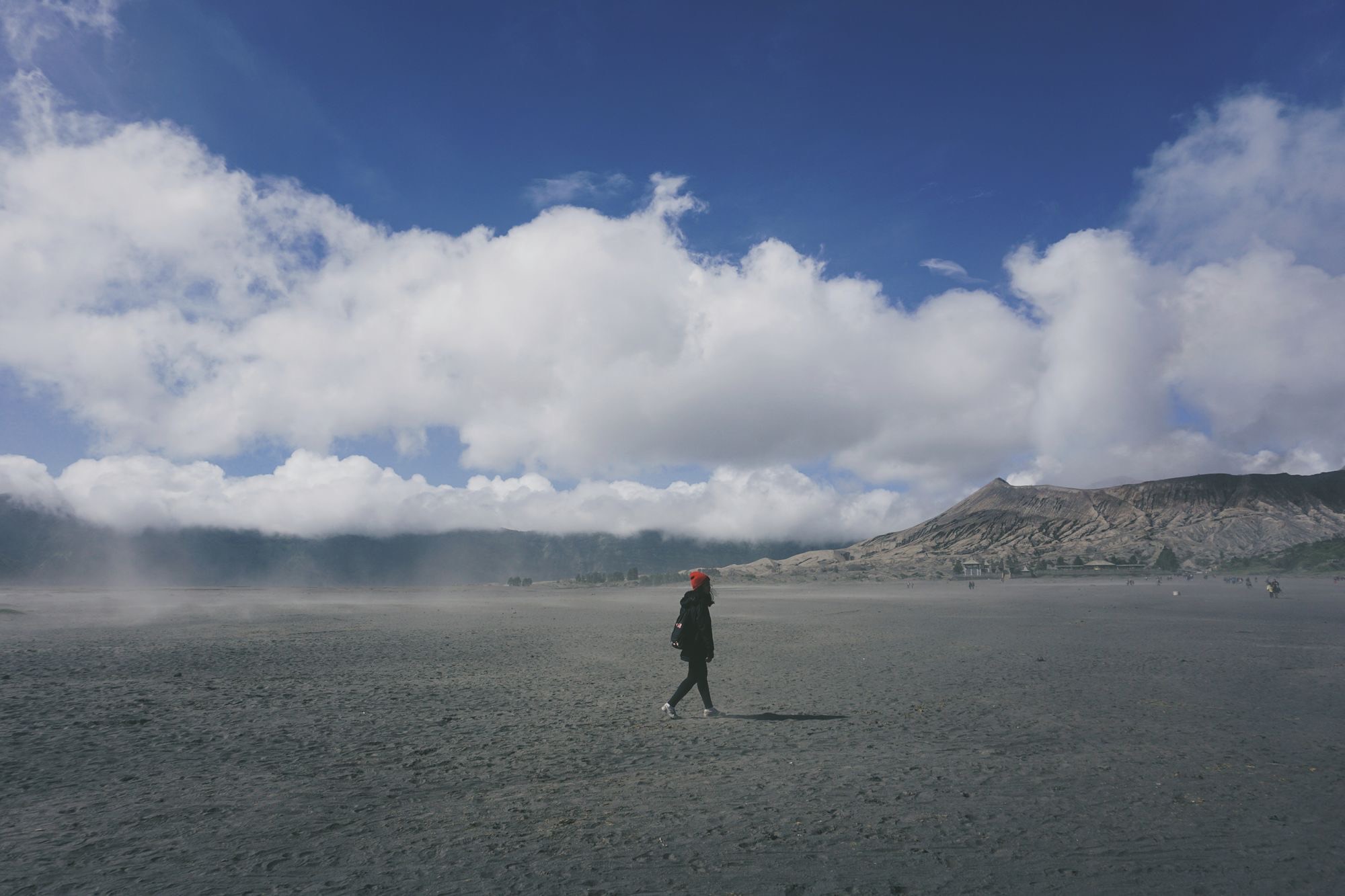 A single person walking across a wide plain of volcanic ash beneath a blue sky.